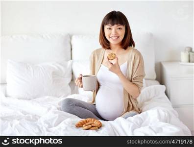 pregnancy, rest, people and breakfast concept - happy pregnant asian woman with cup drinking tea and eating cookie in bed at home bedroom. happy pregnant woman eating cookie in bed at home. happy pregnant woman eating cookie in bed at home