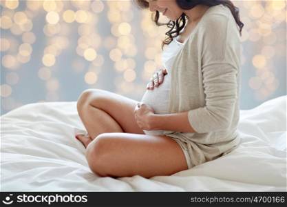 pregnancy, rest, people and expectation concept - close up of happy smiling pregnant woman sitting in bed and touching her belly over holidays lights background