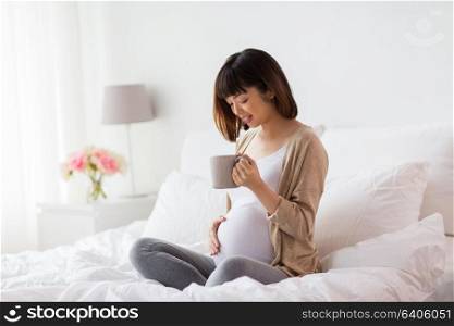 pregnancy, rest, people and expectation concept - happy pregnant asian woman with cup drinking tea in bed at home bedroom. happy pregnant woman with cup drinking tea at home
