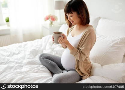 pregnancy, rest, people and expectation concept - happy pregnant asian woman with cup drinking tea in bed at home bedroom. happy pregnant woman with cup drinking tea at home