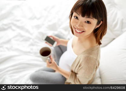 pregnancy, rest, people and expectation concept - happy pregnant asian woman with cup of tea and smartphone in bed at home bedroom. happy pregnant woman with cup of tea in bed