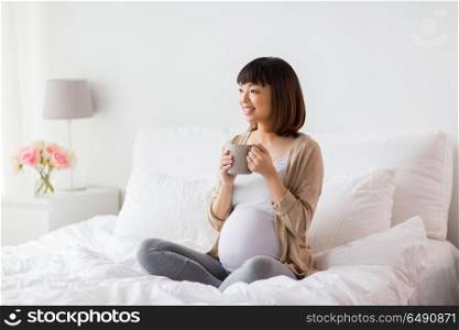 pregnancy, rest, people and expectation concept - happy pregnant asian woman with cup drinking tea in bed at home bedroom. happy pregnant woman with cup drinking tea at home. happy pregnant woman with cup drinking tea at home