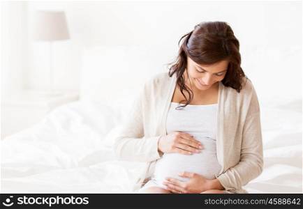 pregnancy, rest, people and expectation concept - happy pregnant woman sitting on bed and touching her belly at home