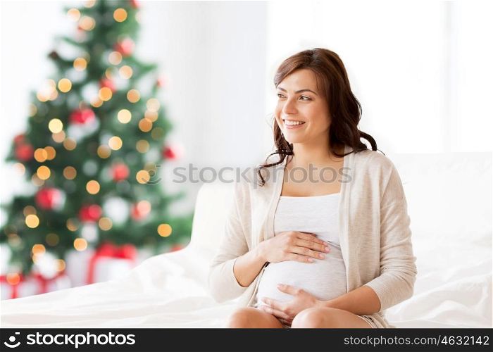 pregnancy, rest, people and expectation concept - happy pregnant woman sitting on bed and touching her belly over christmas tree background