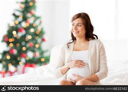pregnancy, rest, people and expectation concept - happy pregnant woman sitting on bed and touching her belly over christmas tree background