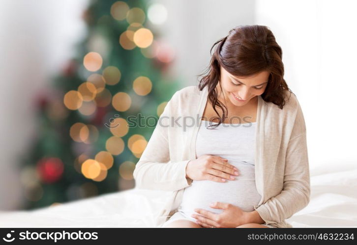 pregnancy, rest, people and expectation concept - happy pregnant woman sitting on bed and touching her belly over christmas tree background