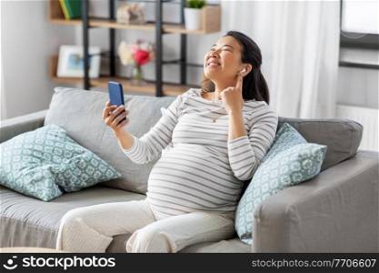 pregnancy, rest, people and expectation concept - happy smiling pregnant asian woman with smartphone and earphones sitting on sofa at home and listening to music. pregnant woman with phone and earphones at home