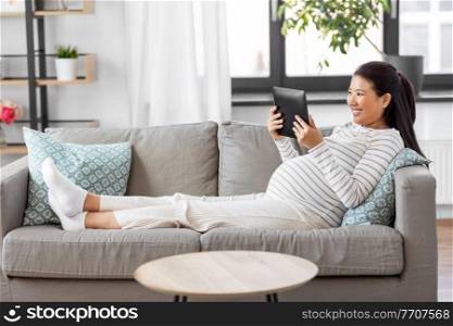 pregnancy, rest, people and expectation concept - happy smiling pregnant asian woman with tablet pc computer sitting on sofa at home. happy pregnant asian woman with tablet pc at home