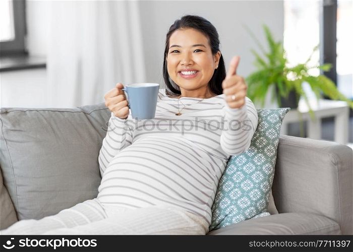 pregnancy, rest, people and expectation concept - happy smiling pregnant asian woman with mug sitting on sofa at home and drinking tea. happy pregnant woman drinking tea at home