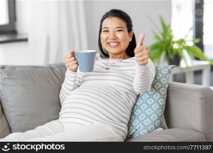 pregnancy, rest, people and expectation concept - happy smiling pregnant asian woman with mug sitting on sofa at home and drinking tea. happy pregnant woman drinking tea at home