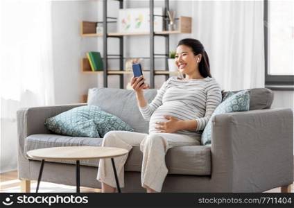 pregnancy, rest, people and expectation concept - happy smiling pregnant asian woman with smartphone and earphones sitting on sofa at home and listening to music. pregnant woman with phone and earphones at home