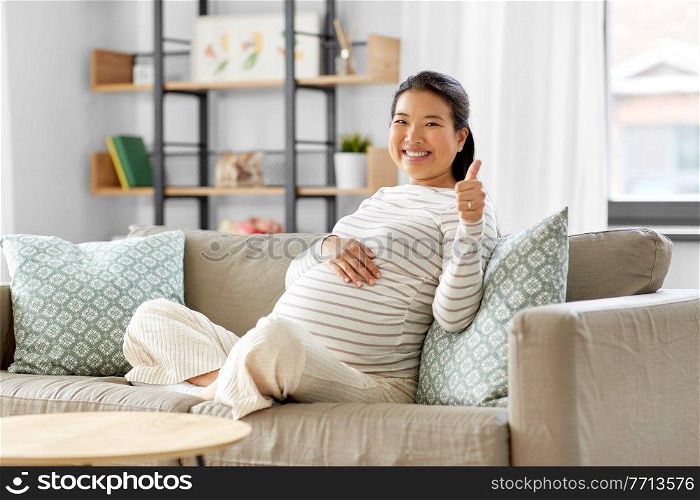pregnancy, rest, people and expectation concept - happy smiling pregnant asian woman sitting on sofa at home and showing thumbs up. happy pregnant asian woman showing thumbs up