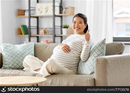 pregnancy, rest, people and expectation concept - happy smiling pregnant asian woman sitting on sofa at home and showing thumbs up. happy pregnant asian woman showing thumbs up