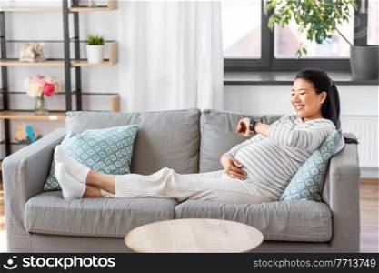 pregnancy, rest, people and expectation concept - happy smiling pregnant asian woman with smart watch sitting on sofa at home. happy pregnant woman with smart watch at home