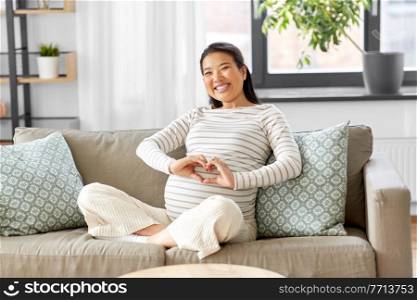 pregnancy, rest, people and expectation concept - happy smiling pregnant asian woman showing hand heart gesture sitting on sofa at home. happy pregnant asian woman showing heart gesture