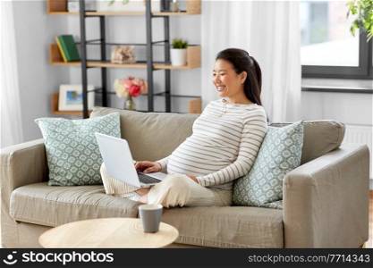 pregnancy, rest, people and expectation concept - happy smiling pregnant asian woman with laptop computer sitting on sofa at home. happy pregnant asian woman with laptop at home