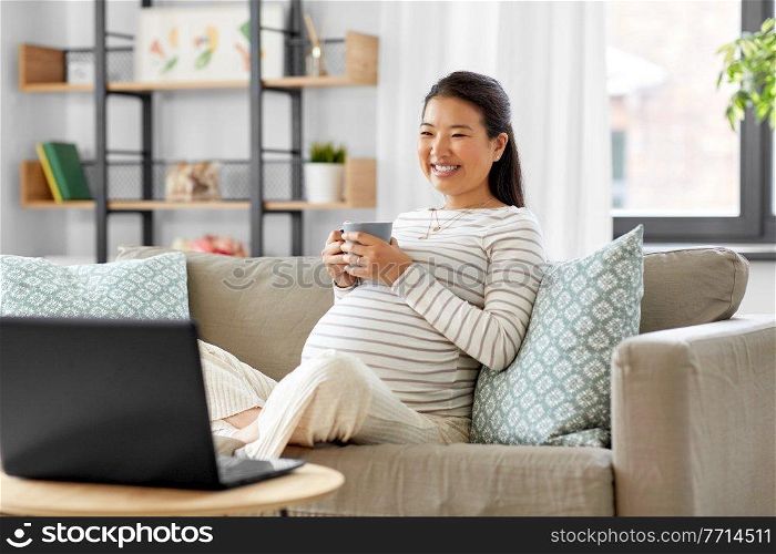 pregnancy, rest, people and expectation concept - happy smiling pregnant asian woman with laptop computer sitting on sofa at home and drinking tea. happy pregnant asian woman with laptop at home