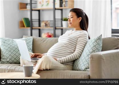 pregnancy, rest, people and expectation concept - pregnant asian woman with laptop computer sitting on sofa at home. pregnant asian woman with laptop at home