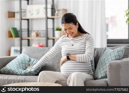 pregnancy, rest, people and expectation concept - pregnant asian woman with smart watch sitting on sofa at home having labor contractions pain. pregnant woman having labor contractions at home