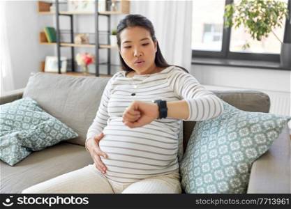 pregnancy, rest, people and expectation concept - pregnant asian woman with smart watch having labor contractions sitting on sofa at home and breathing. pregnant woman having labor contractions at home