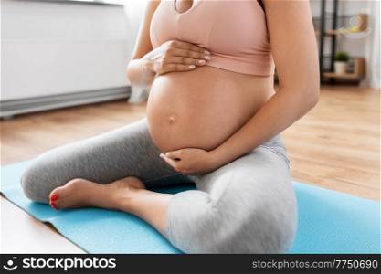 pregnancy, sport and fitness concept - close up of pregnant woman sitting on yoga mat at home and touching her belly. close up of pregnant woman doing yoga at home