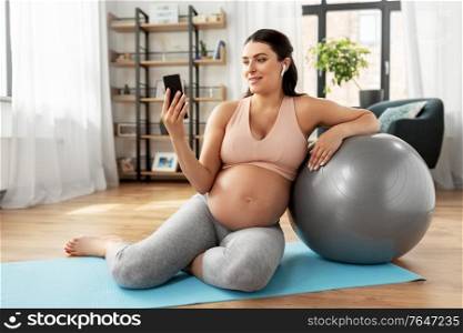 pregnancy, sport and fitness concept - happy smiling pregnant woman with smartphone, earphones and fitball exercising at home. pregnant woman with smartphone and fitball at home