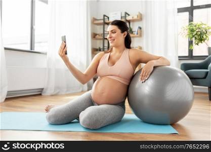 pregnancy, sport and fitness concept - happy smiling pregnant woman with smartphone and fitball exercising at home. pregnant woman with smartphone and fitball at home