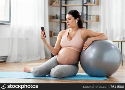 pregnancy, sport and people concept - happy pregnant woman with smartphone, earphones and fitness ball at home. pregnant woman with phone and fitness ball at home