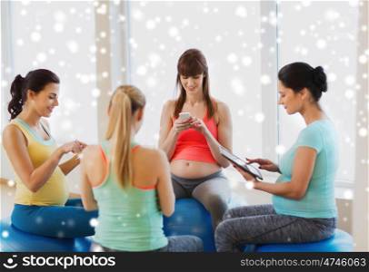pregnancy, sport, fitness, people and healthy lifestyle concept - group of happy pregnant women with tablet pc computer and smartphone sitting on stability balls in gym over snow