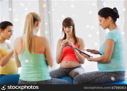 pregnancy, sport, fitness, people and healthy lifestyle concept - group of happy pregnant women with tablet pc computer and smartphone sitting on balls in gym over snow