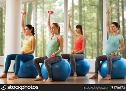 pregnancy, sport, fitness, people and healthy lifestyle concept - group of happy pregnant women with dumbbells exercising on ball in gym