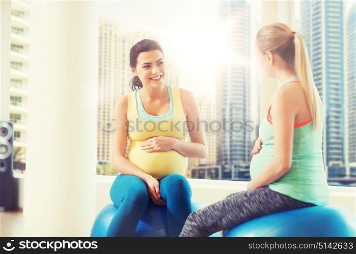 pregnancy, sport, fitness, people and healthy lifestyle concept - two happy pregnant women sitting and talking on balls in gym over city window view background. two happy pregnant women sitting on balls in gym