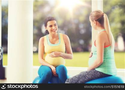 pregnancy, sport, fitness, people and healthy lifestyle concept - two happy pregnant women sitting and talking on balls in gym over natural window view background. two happy pregnant women sitting on balls in gym