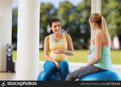 pregnancy, sport, fitness, people and healthy lifestyle concept - two happy pregnant women sitting and talking on balls in gym over natural window view background