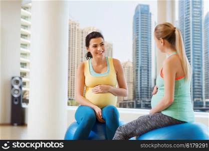 pregnancy, sport, fitness, people and healthy lifestyle concept - two happy pregnant women sitting and talking on balls in gym over city window view background