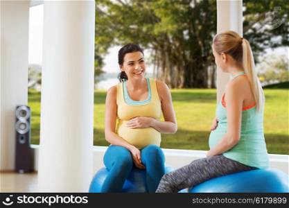 pregnancy, sport, fitness, people and healthy lifestyle concept - two happy pregnant women sitting and talking on balls in gym over natural window view background