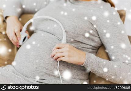 pregnancy, technology and people concept - close up of pregnant woman with headphones on her belly at home over snow. pregnant woman with headphones listening to music