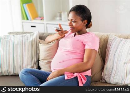 pregnancy, technology and people concept - happy pregnant african american woman using voice command recorder or calling on smartphone at home. pregnant woman using voice recorder on smartphone