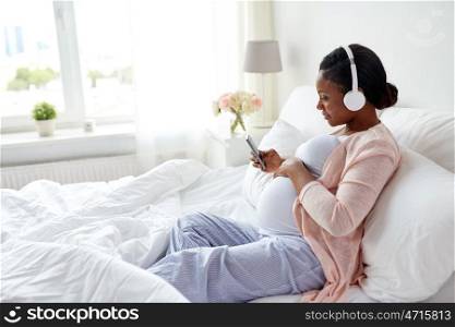 pregnancy, technology and people concept - happy pregnant african american woman with headphones and smartphone in bed at home. pregnant woman in headphones with smartphone. pregnant woman in headphones with smartphone