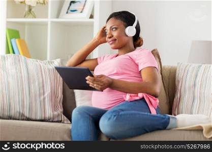 pregnancy, technology and people concept - happy pregnant african american woman with headphones and tablet pc computer at home. pregnant woman in headphones with tablet pc