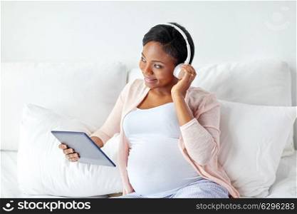 pregnancy, technology and people concept - happy pregnant african american woman with headphones and tablet pc computer in bed at home. pregnant woman in headphones with tablet pc