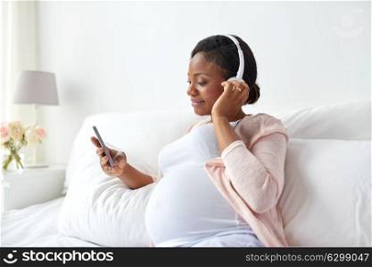 pregnancy, technology and people concept - happy pregnant african american woman with headphones and smartphone in bed at home. pregnant woman in headphones with smartphone