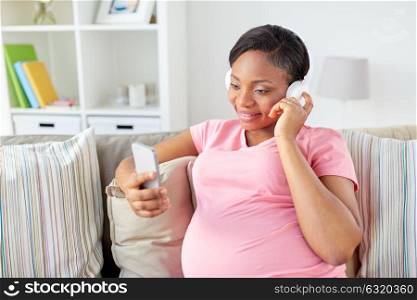pregnancy, technology and people concept - happy pregnant african american woman with headphones and smartphone listening to music at home. pregnant woman in headphones with smartphone