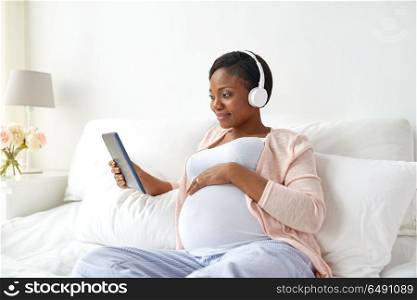 pregnancy, technology and people concept - happy pregnant african american woman with headphones and tablet pc computer in bed at home. pregnant woman in headphones with tablet pc. pregnant woman in headphones with tablet pc