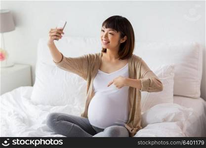 pregnancy, technology and people concept - happy pregnant asian woman taking selfie by smartphone in bed at home and pointing finger to her belly. pregnant woman taking selfie by smartphone at home