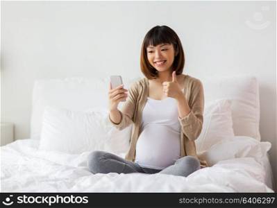 pregnancy, technology and people concept - happy pregnant asian woman taking selfie by smartphone in bed at home and showing thumbs up. pregnant woman taking selfie by smartphone at home