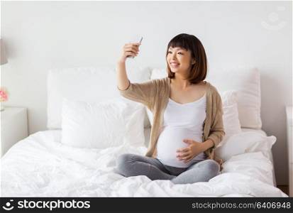 pregnancy, technology and people concept - happy pregnant asian woman taking selfie by smartphone in bed at home. pregnant woman taking selfie by smartphone at home