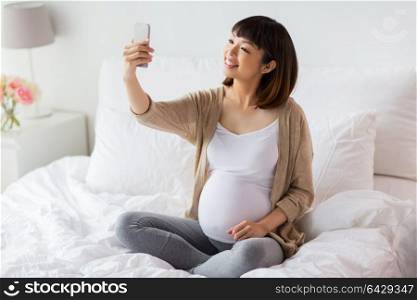 pregnancy, technology and people concept - happy pregnant asian woman taking selfie by smartphone in bed at home. pregnant woman taking selfie by smartphone at home