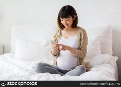pregnancy, technology and people concept - happy pregnant asian woman with smartphone in bed at home. pregnant woman with smartphone in bed at home