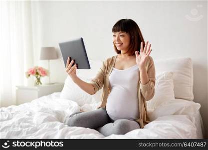 pregnancy, technology and people concept - happy pregnant asian woman with tablet pc computer having video call and waving hand in bed at home. happy pregnant asian woman with tablet pc at home